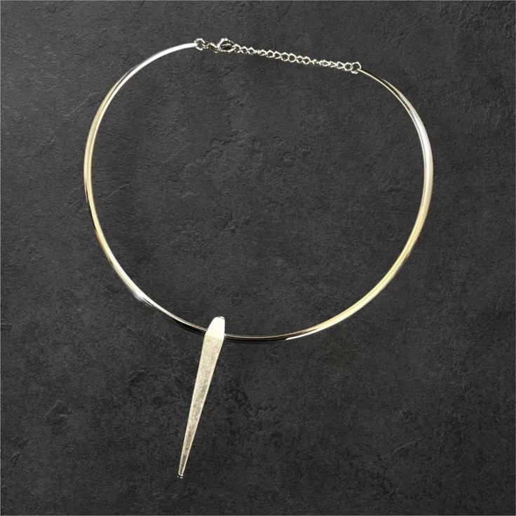 a silver necklace with a thin, rounded choker and a long, pointed pendant hangs on a dark background. the necklace is simple and modern in design and is a great statement piece.