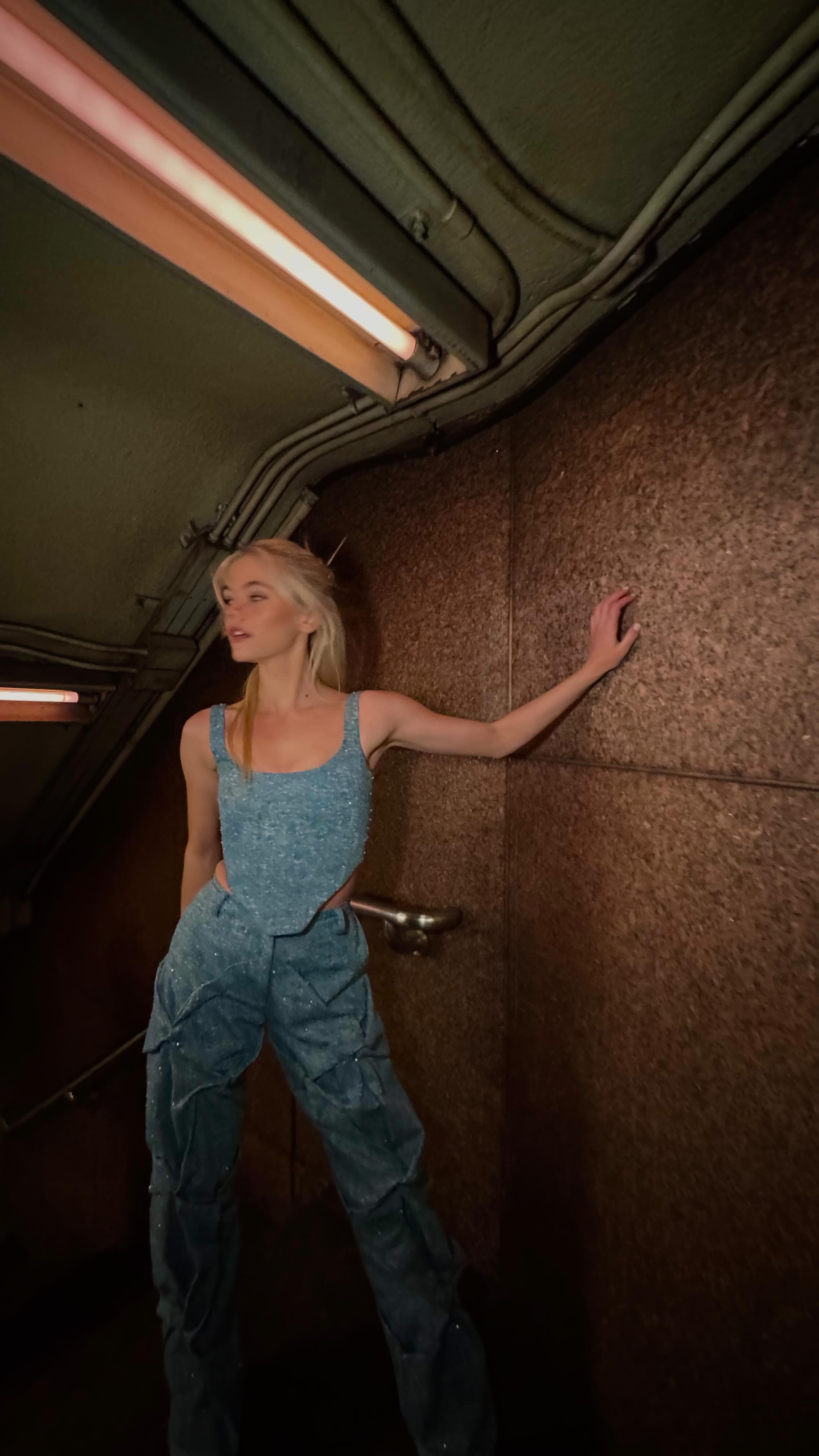 a blonde woman in a denimlike tank top and matching pants is standing in what appears to be a subway tunnel. she is leaning slightly against a textured wall with her hand resting on it. above her, a fluorescent light fixture illuminates the scene. the overall setting suggests an underground or industrial environment.
