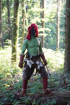 a person in cosplay, dressed as a greenskinned orc with long red hair, stands in a forest.  the orc is wearing a costume that includes a tattered brown outfit, maroon leggings, and carries what appears to be an axe. the scene is set in a lush woodland environment with dappled sunlight filtering through the trees. the cosplay is detailed and appears wellexecuted.