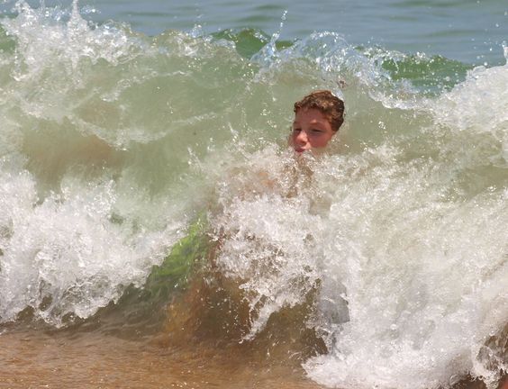 a young boy with short brown hair is being covered by a large wave in the ocean. the white foam of the wave surrounds him, and his face is partially visible. the background shows more ocean water, and the foreground shows a glimpse of the sandy beach.