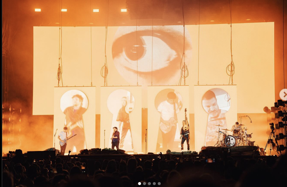 a band is performing on stage at a concert. the stage is lit up with bright lights. the band is playing guitars and drums. the audience is cheering and applauding. there are  keyhole and eye shaped images on the screen behind the band.