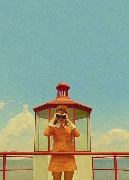 a young woman wearing a bright orange dress and a white collar stands in front of a red and white lighthouse. she is looking through binoculars and her expression is intense. the image has a vintage feel and the colors are bright and bold. the sky is blue and there are clouds in the background.  the woman is looking out at the ocean.