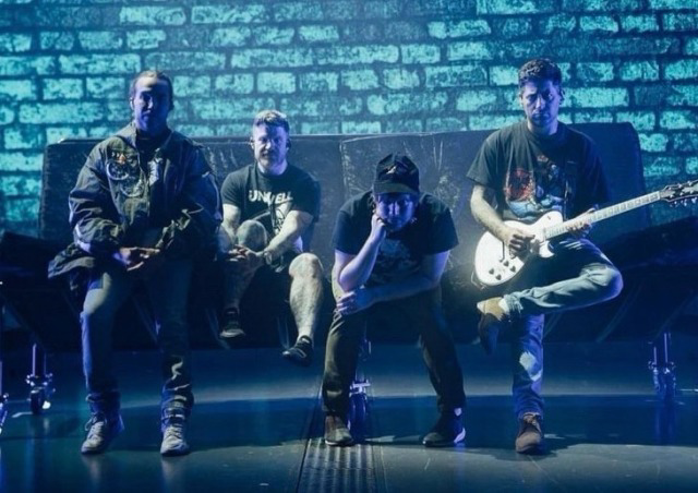 four men in a rock band are performing on a stage with blue lights. the man on the far right is playing an electric guitar. the man in the middle is looking down and has his hand on his chin. the man on the far left is looking off into the distance and the man in the middle left is sitting with his feet crossed, looking at the ground.