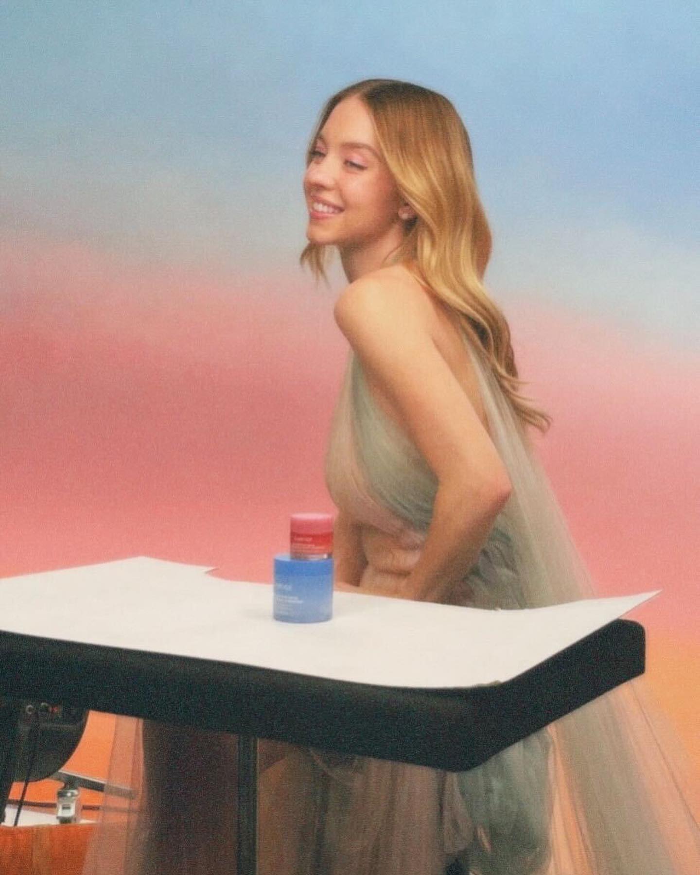 the image shows a young, smiling woman, possibly a model or influencer, wearing a light teal, flowing tulle dress. she is positioned slightly turned away from the camera, looking over her shoulder.  she's standing in front of a pink and blue gradient backdrop. on a small table in front of her are two small containers, one pink and one blue, which appear to be skincare or cosmetic products. the overall setting suggests a professional photoshoot or advertisement for beauty products. the lighting is soft and the mood is light and airy.