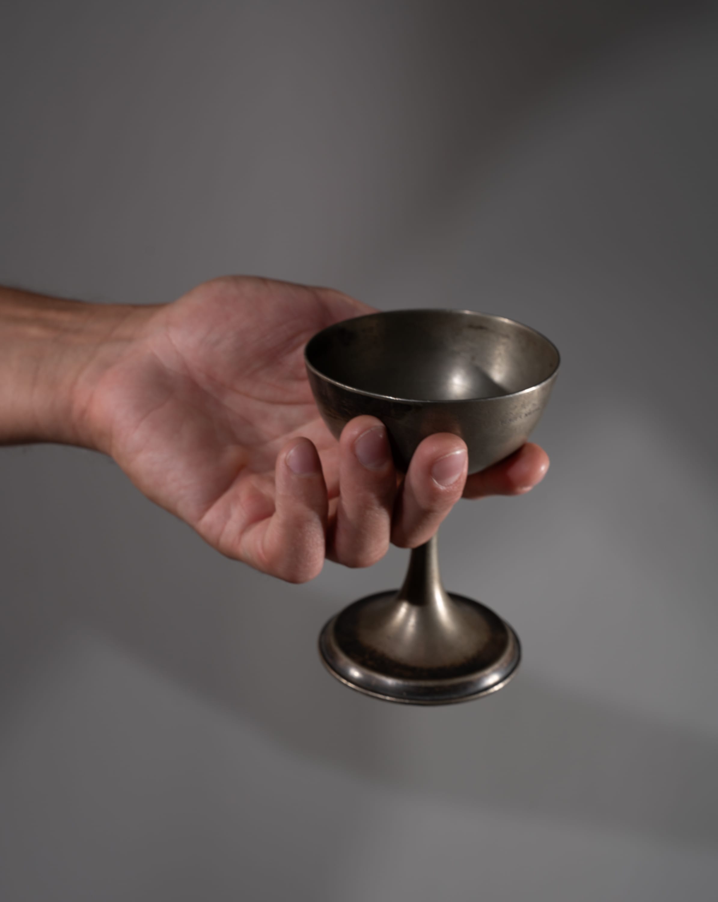 a man's hand holds a vintage silver cup against a neutral gray background. the cup is made of tarnished metal and has a simple design with a rounded bowl and a slender stem. the lighting is soft and even, highlighting the texture of the metal and the details of the man's hand.