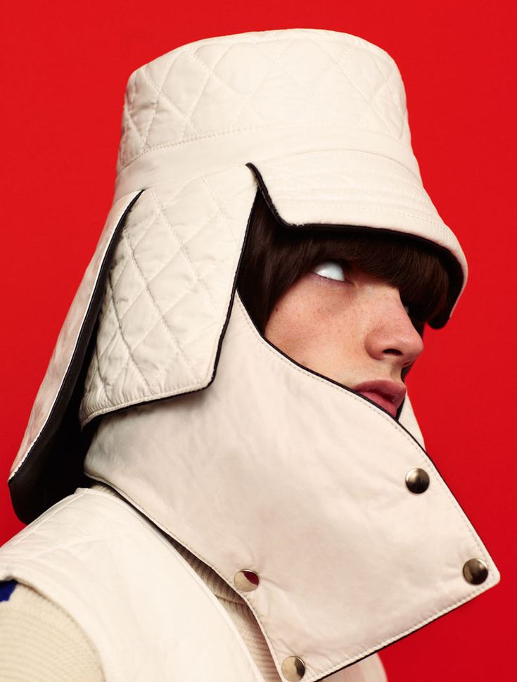 a man in a white quilted hat and jacket looks up at something off camera. he has a black trim on his hat and coat and the image is shot against a red background.  the hat is pulled down low on his forehead and the brim of the hat blocks much of his hair from view. the man is looking up and the expression on his face is calm. his eyes are open and his mouth is slightly open.