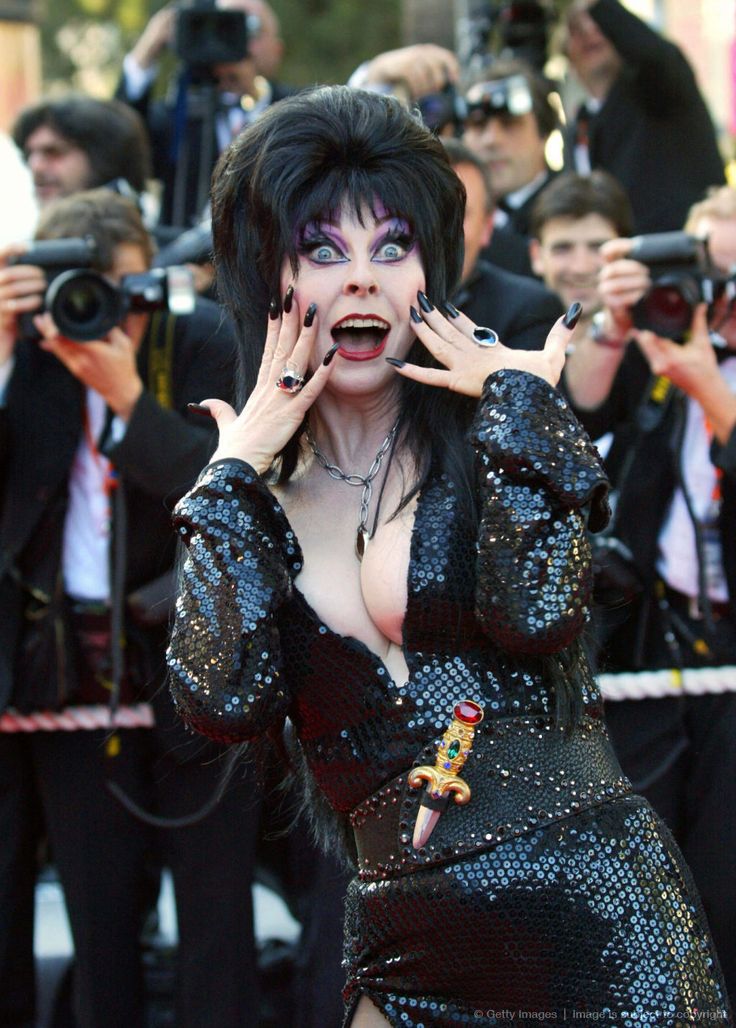 a woman with long dark hair, wearing a black sequined dress and a dagger belt, smiles and appears shocked or surprised as she is photographed by paparazzi on the red carpet. she has a large smile on her face and is holding her hands up in a surprised gesture.