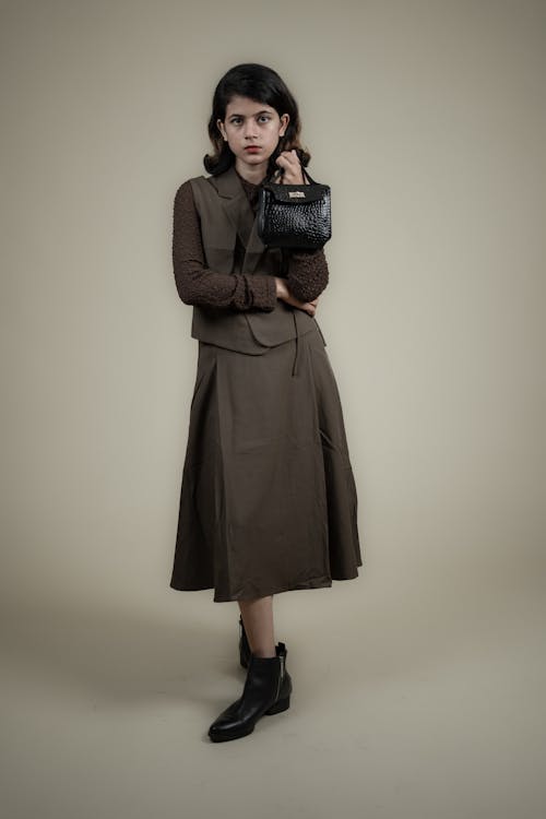 a full shot of a young woman in a fashionable outfit standing in a studio setting against a neutral background. she is wearing a brown skirt that reaches midcalf, paired with a matching brown vest. underneath the vest, she has a longsleeved top with a textured pattern. the outfit is completed with black ankle boots. she is holding a small, black, textured handbag in one hand. her expression is neutral as she looks directly at the camera. the lighting is soft, highlighting the textures of the clothing and the details of the accessories.