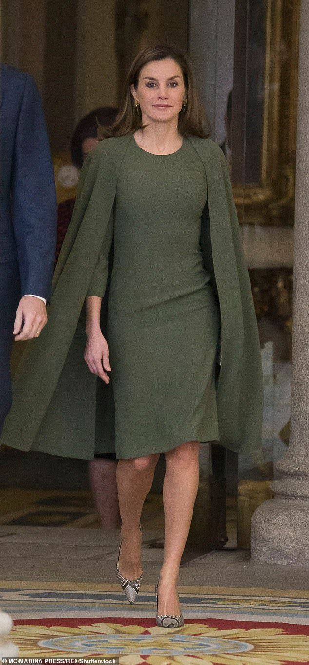 queen letizia of spain is captured in a fulllength shot, walking towards the camera. she's elegantly dressed in a kneelength olive green dress paired with a matching cape.  her outfit is complemented by sophisticated silver heels.  the setting appears to be a grand palace entrance, suggesting a formal event or official engagement. the background is subtly blurred, focusing attention on the queen. a portion of another person's suit is visible in the left foreground, while another individual can be partially seen behind her. the ornate carpet underfoot suggests a grand interior. the overall image projects sophistication and royal presence.