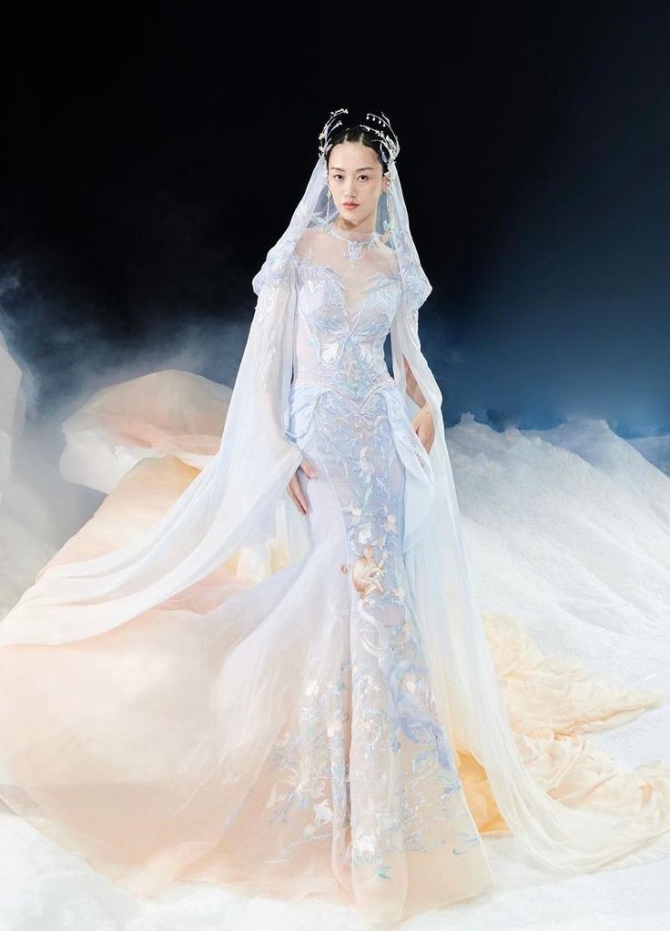 a model wearing a stunning white gown with delicate blue embroidery and a long veil, poses gracefully on a runway. the dress has a fitted bodice and a flowing skirt, creating an ethereal and elegant look. her hair is styled with a sparkling crown and she looks confident and beautiful. the gown is perfect for a bride seeking a romantic and timeless look.