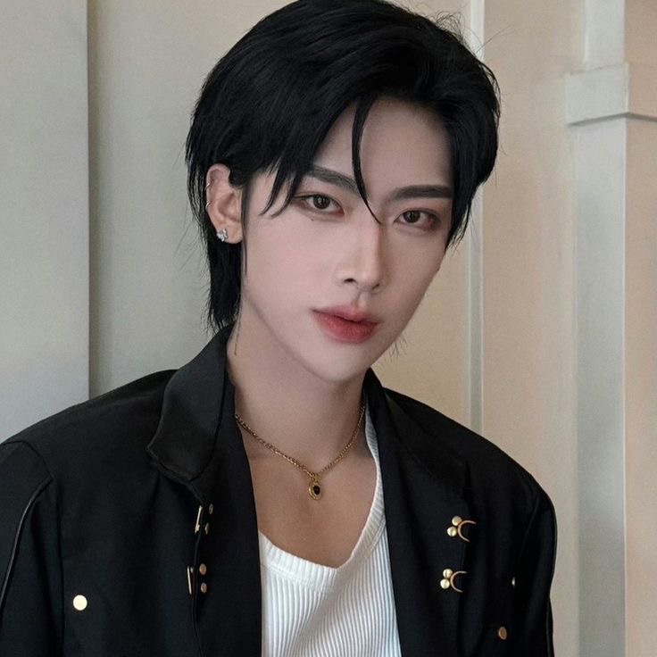 a young asian man with black hair styled with a middle part poses for the camera. he is wearing a black jacket over a white shirt, and a gold chain necklace. he is lit with soft lighting and the overall image has a warm tone.