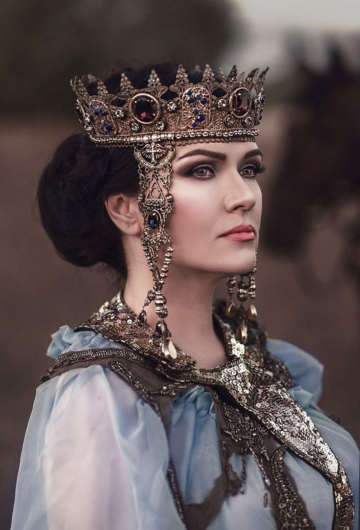 woman is captured in a stunning portrait, adorned in a majestic, ornate gold crown and elaborate jewelry.  the crown is richly decorated with deepcolored gems, and the overall aesthetic suggests a regal, possibly fantasy or historical, setting.  the woman's expression is serious and contemplative, enhancing the image's sense of grandeur and power. her light blue, sheer gown complements the jewelry's shimmer and the crown's metallic sheen. the background is softly blurred, drawing focus to the intricate details of her attire and regal bearing.