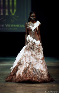 a black woman model walks the runway wearing a beautiful unique gown made of white and brown fabric.