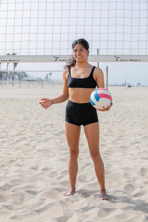 a young hispanic woman, smiling, stands on a sandy beach volleyball court in santa monica, california.  she's wearing a black bikini top and shorts and holds a volleyball.  the volleyball net is clearly visible in the background.  the scene is bright and sunny, depicting a summer day.  the overall impression is of an active, healthy lifestyle and a fun beach volleyball game.