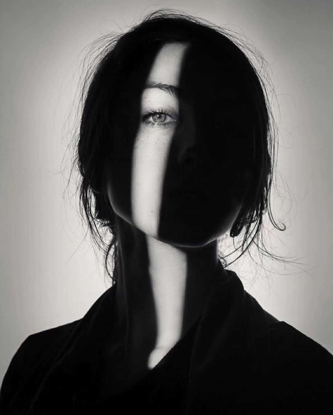 a black and white portrait of a woman with her face half in shadow. the light coming from behind her casts lines of light across her face and neck. she has dark hair and a mysterious expression. the image is very dramatic and moody.
