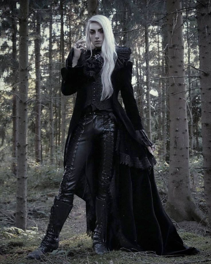 a pale man with long white hair, wearing dark gothic attire including a long coat, vest, leather pants, and boots, stands in a forest. he is adorned with rings and makeup, and the clothing features frills, ruffles, and lace. the scene has a mysterious and dark fantasy aesthetic.