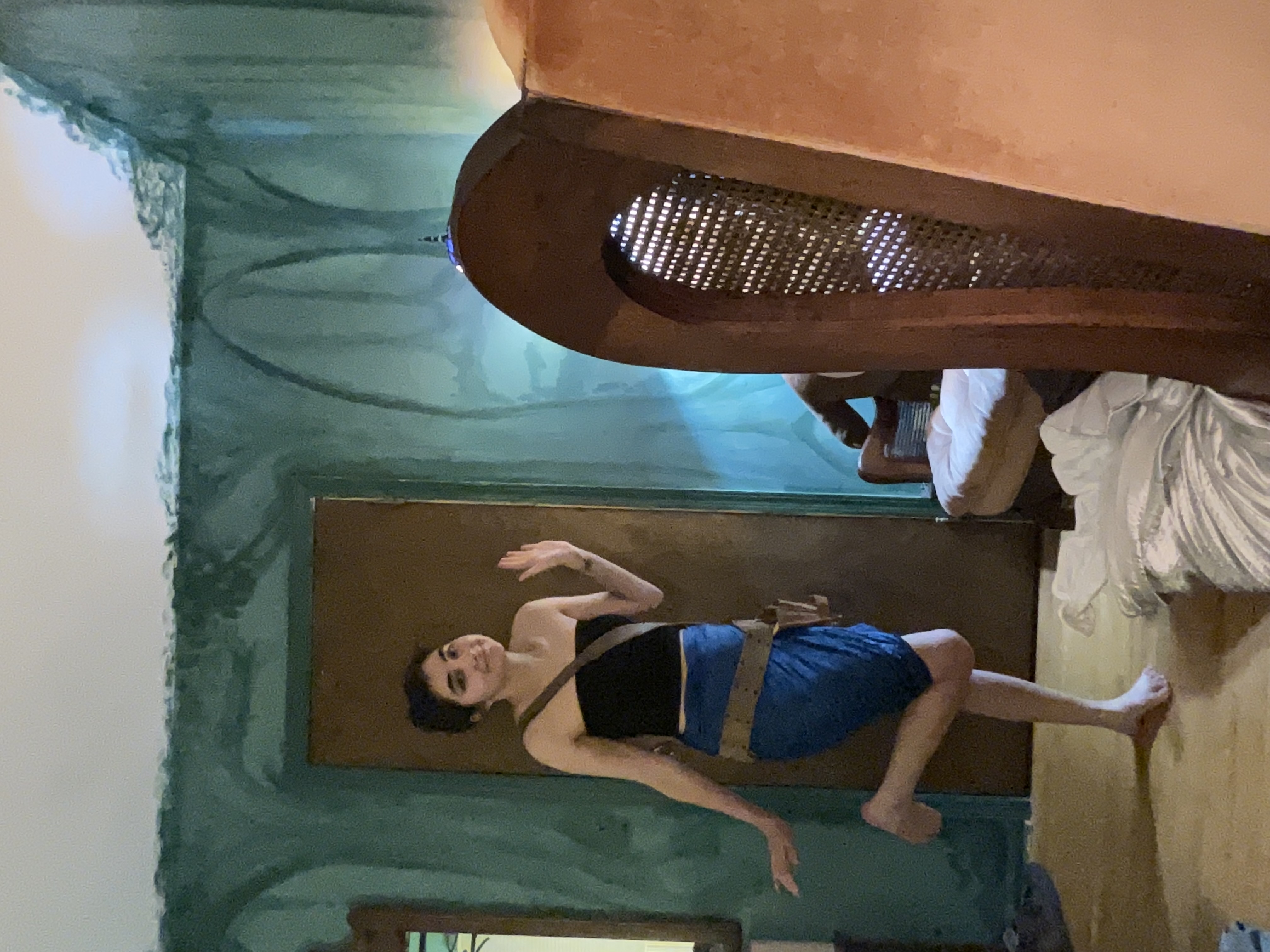 a young woman with dark hair stands in a room with teal walls that are unevenly painted. she's wearing a blue wrap skirt and a black bandeau top.  she is posing with one leg slightly bent.  in the background, there's a partially visible person sitting in a chair and a piece of vintage furniture with a cane back. the room appears to be undergoing renovation or is in the midst of a home project. the overall feel is casual and slightly messy.