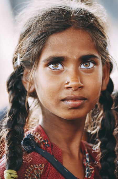a portrait of a young girl with dark skin, striking lightcolored eyes, and braided hair. she has a nose piercing and is wearing a patterned red shirt. her expression is serious and direct.