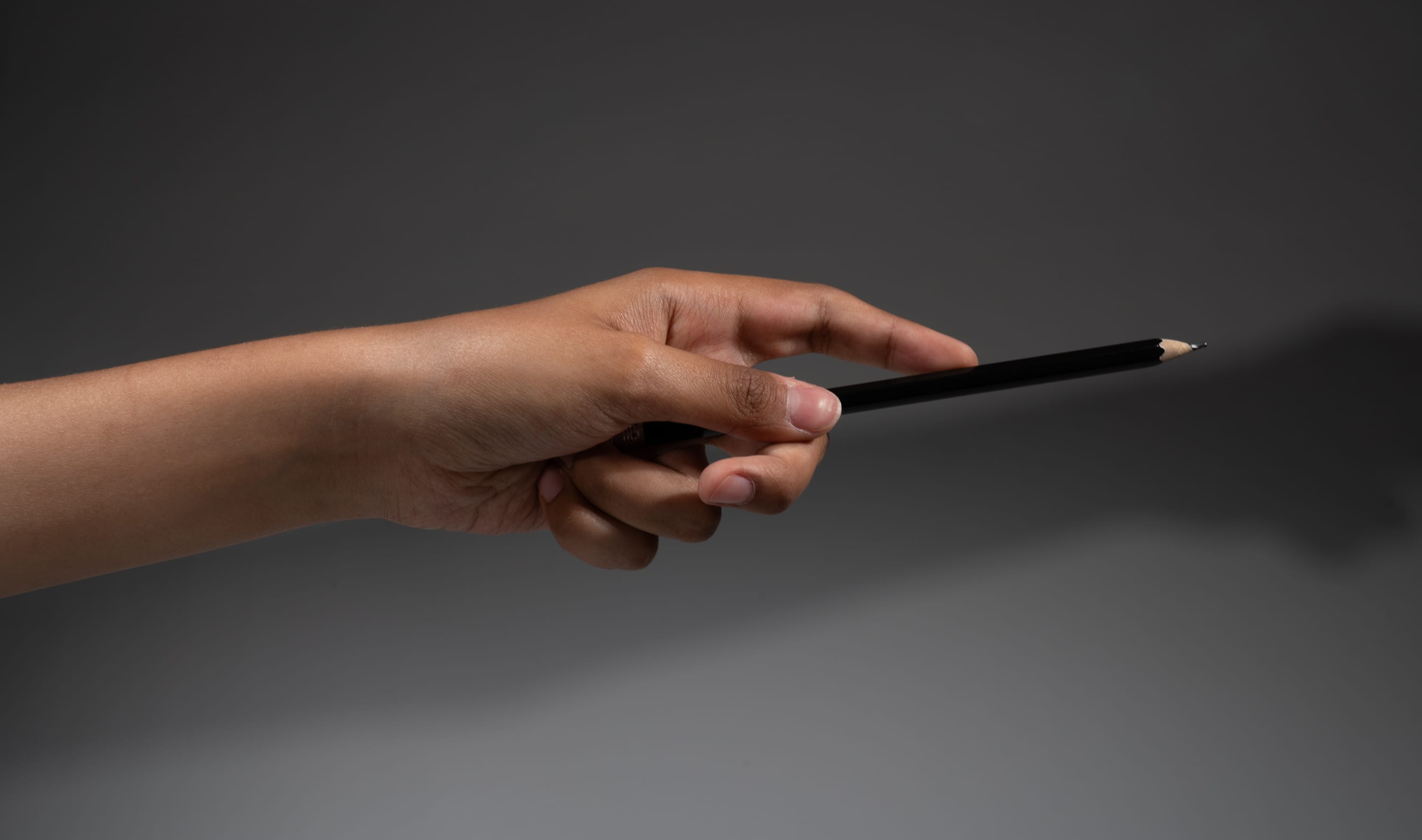 a hand is holding a black pencil and pointing towards the right side of the image against a dark gray background. a shadow of the hand and pencil is cast on the background.