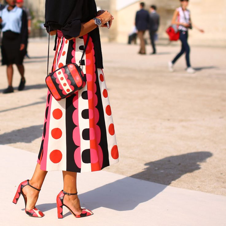 a woman is captured from the knees down in this street style shot, showcasing her fashionable outfit.  she's wearing a midi skirt with a bold geometric print featuring red, pink, black, and white polka dots and circles. the skirt is paired with a black top and bright red/pink block heel ankle strap shoes. she carries a small, stylish crossbody bag in a complementary color scheme. the background is blurred, indicating an outdoor location, likely a city street, with other people visible in the distance but out of focus. the overall image evokes a sense of chic and effortless parisian style.