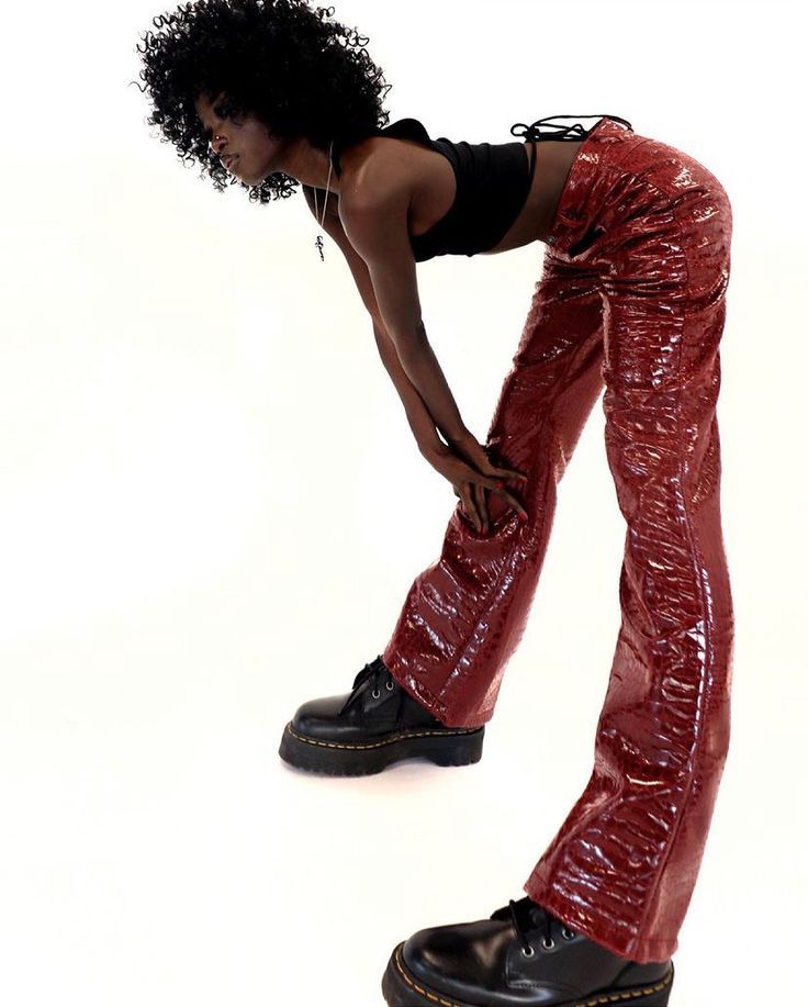 a young woman with dark curly hair is posing against a white background. she is wearing a red pair of leather flare pants with a black crop top. she has on a black necklace and black boots. she is standing in a stylish pose.