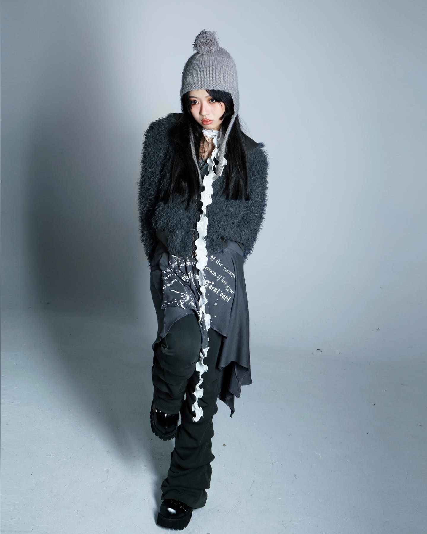 a young woman is posing in a studio setting against a plain light grey backdrop. she is wearing a grey fuzzy jacket, a long grey scarf, dark pants and boots, and a matching grey beanie. the outfit is stylish and somewhat edgy and has a cool vibe. the model's expression is somewhat serious and looking downwards.