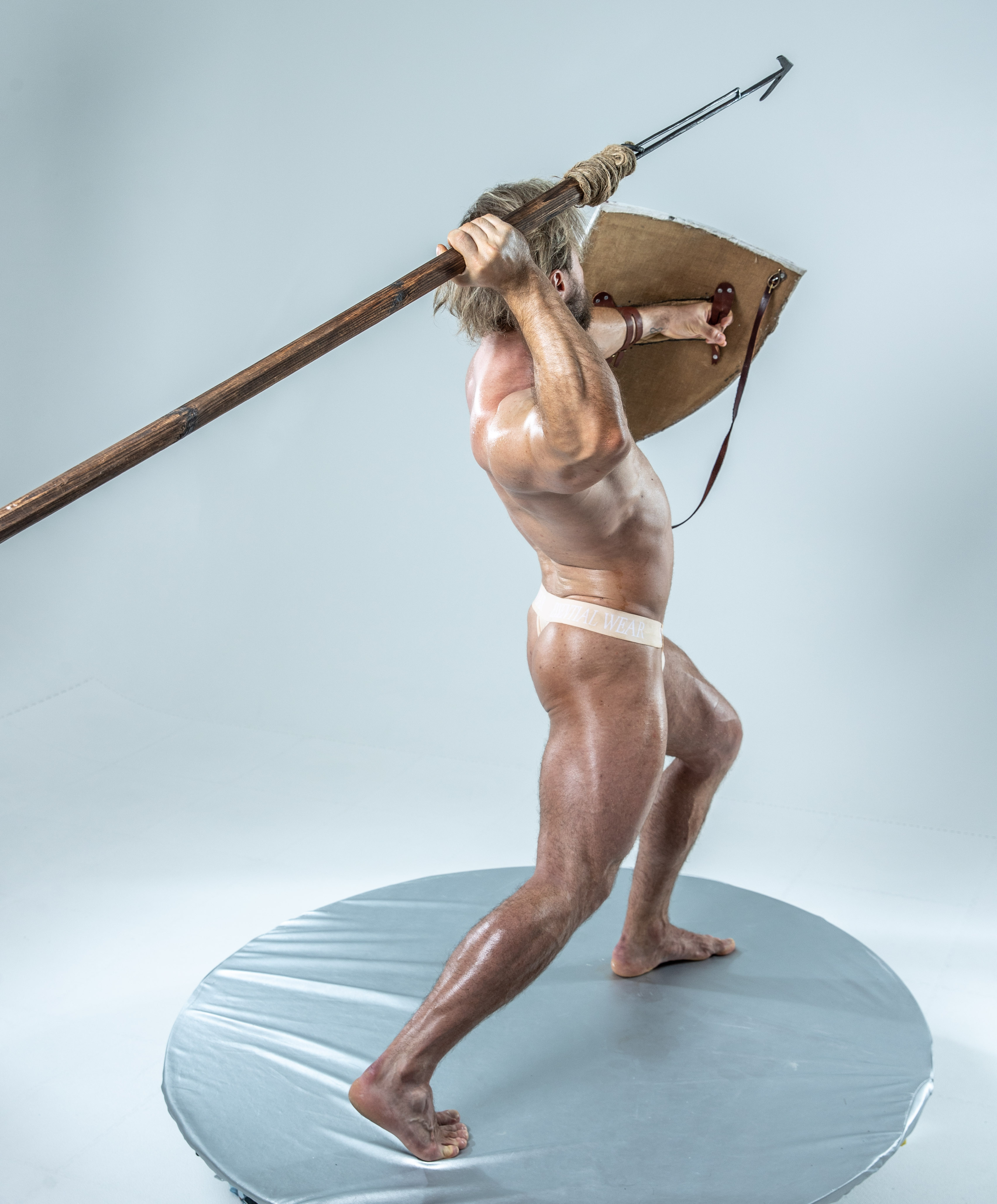 a muscular man with gray hair and a white loincloth stands on a gray platform, wielding a spear and shield. he is positioned in a dynamic pose, suggesting combat. the scene is brightly lit in a studio setting.