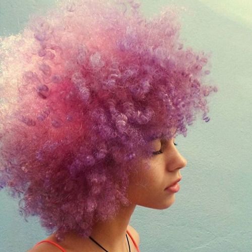 a young woman with vibrant purple and pink afro hair smiles as she gazes off to the side.