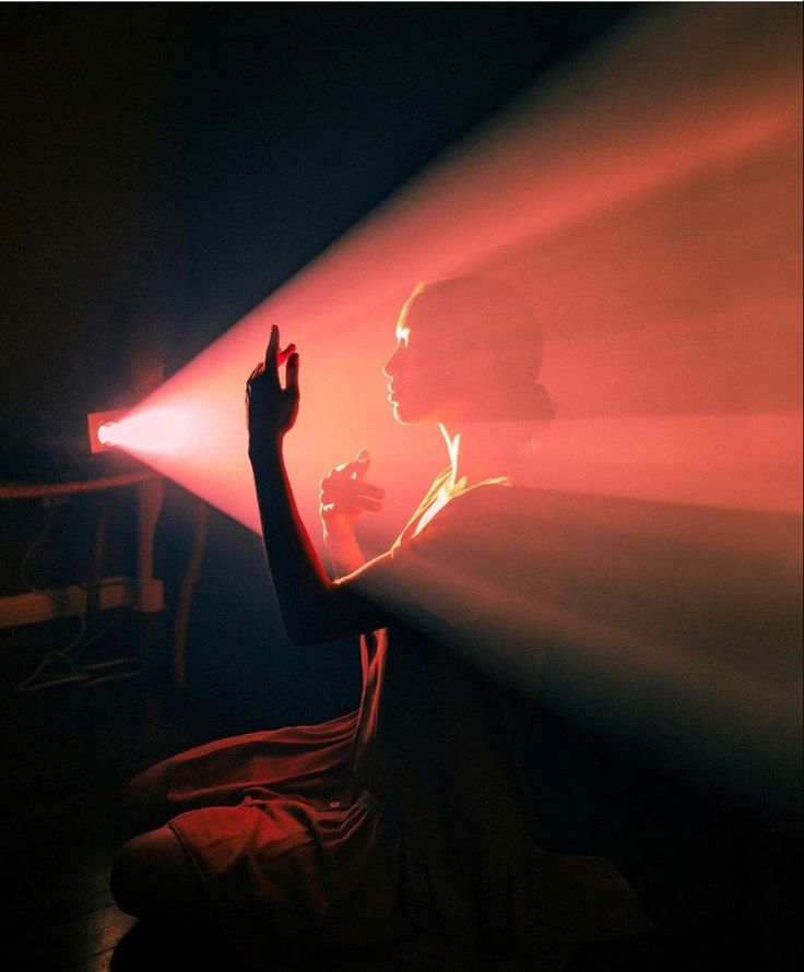 a silhouette of a woman standing in a dark room. she is illuminated by a red spotlight that casts a dramatic shadow on the wall behind her. her right hand is raised, as if she is reaching out to something. the image is mysterious and cinematic.
