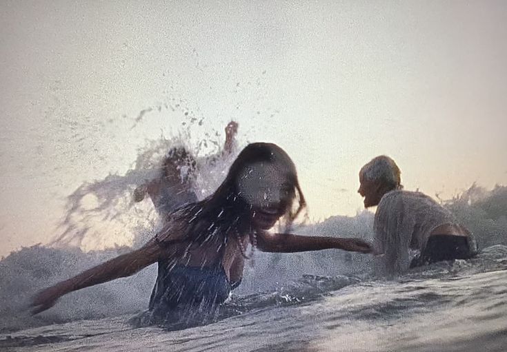 the image captures a dynamic moment of several young adults joyfully splashing in ocean waves. the setting appears to be a beach at either dusk or dawn, with soft, muted lighting.  the focus is on a young woman with long dark hair in the center foreground, her face partially obscured by water and movement.  others are around her, their forms less distinct due to the splashing and the photographic style. the overall mood is one of carefree fun and youthful energy. the image has a slightly grainy or filmlike quality with noticeable motion blur from the water's movement.