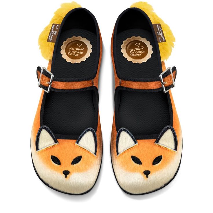 an overhead shot shows a pair of hot chocolate design mary jane shoes decorated to look like the face of an orange and white fox. the shoes have rounded toes that feature the lower half of the fox's white face, black nose, and black angled eyes. orange "fur" covers the rest of the toe, up to the black opening and strap. a buckle closure secures the orange strap to the side of the shoe. behind each shoe, yellow faux fur can be seen. the insole has the hot chocolate design logo.