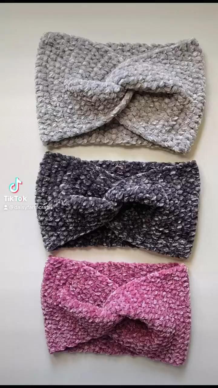 three crocheted headbands are displayed in a vertical stack on a white surface. the headbands have a twisted design in the center and are made of velvet yarn. the top headband is gray, the middle one is dark gray, and the bottom one is pink. the headbands appear to be handmade.