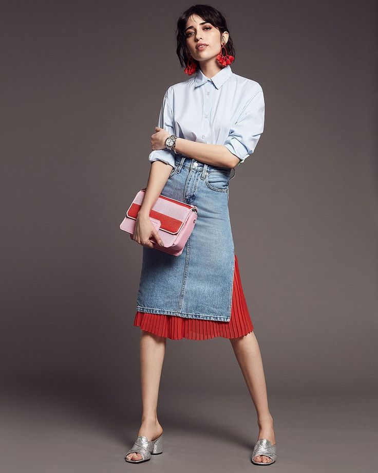 a woman model is posing in a studio with a gray background. she is wearing a white button down shirt, blue denim skirt with a red pleated fabric underneath, and a pink and red bag. she is also wearing silver shoes, and red earrings. she is looking at the camera.
