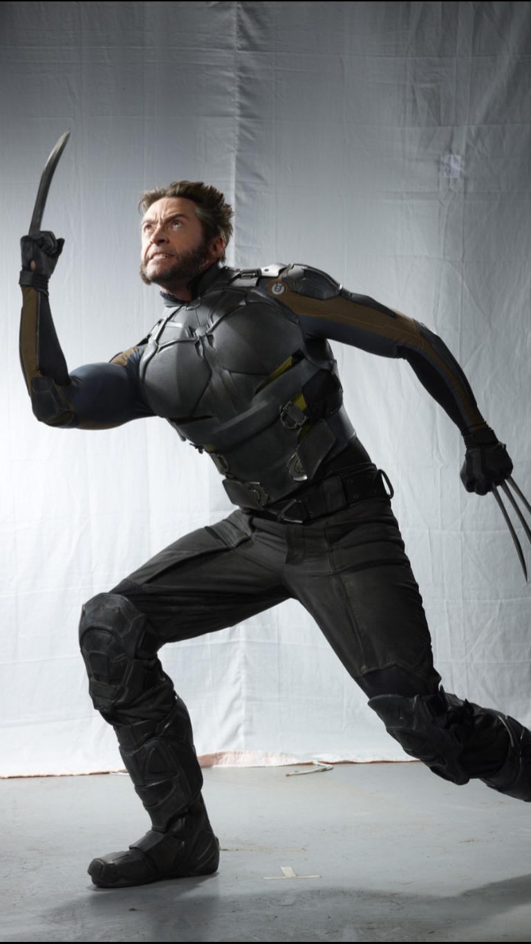 a full shot of hugh jackman dressed as wolverine in a black and grey costume with metal claws extended from one hand. he is in an action pose.