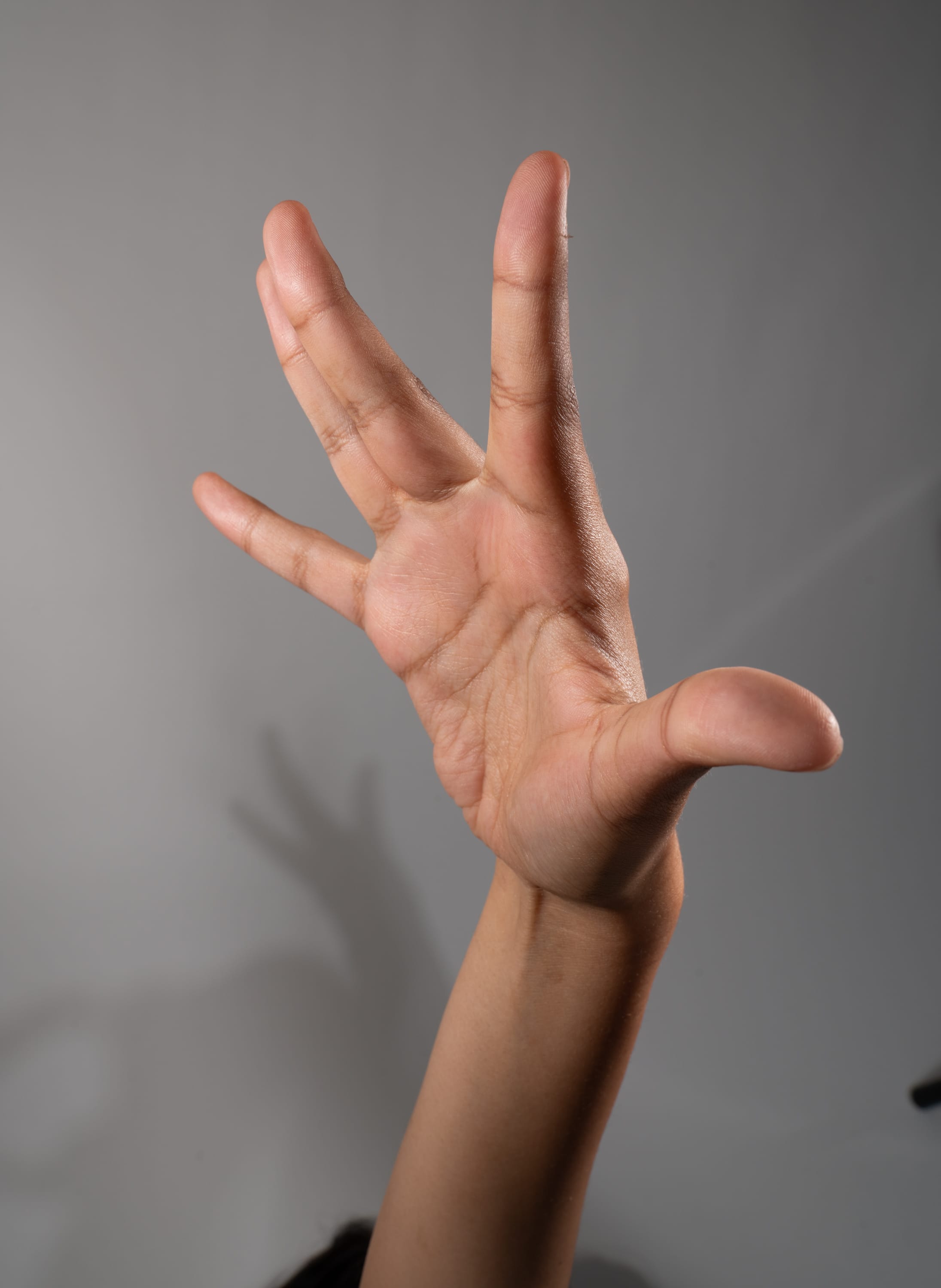 a closeup image of a person's hand extended upwards against a gray background. the hand is positioned with the fingers spread apart, creating a dynamic and expressive gesture. the studio lighting highlights the contours and textures of the skin, revealing the intricate details of the human hand.