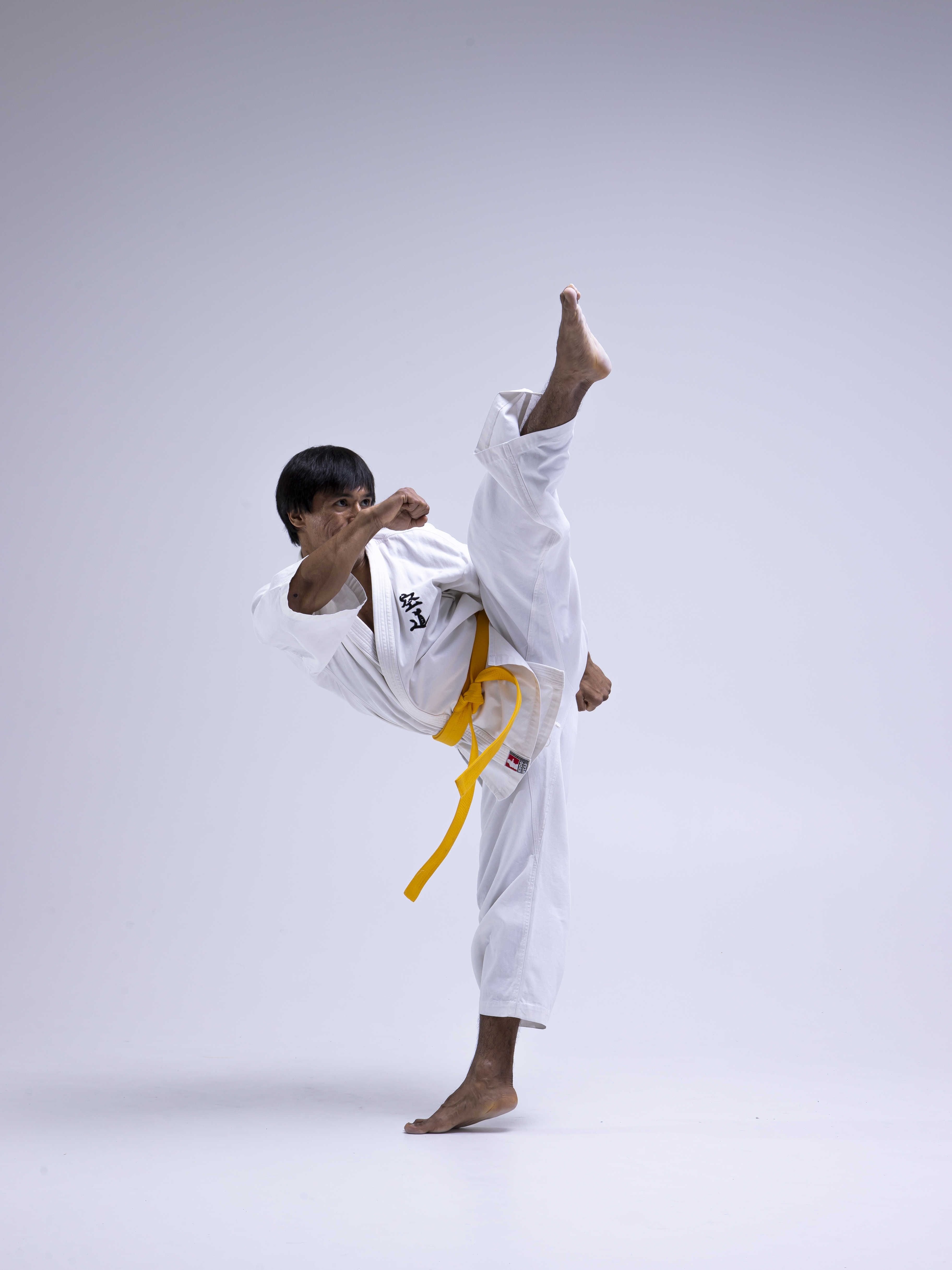 a man in a white karate uniform with a yellow belt performs a high kick in a martial arts stance. he is barefoot and the background is a plain light gray or white.