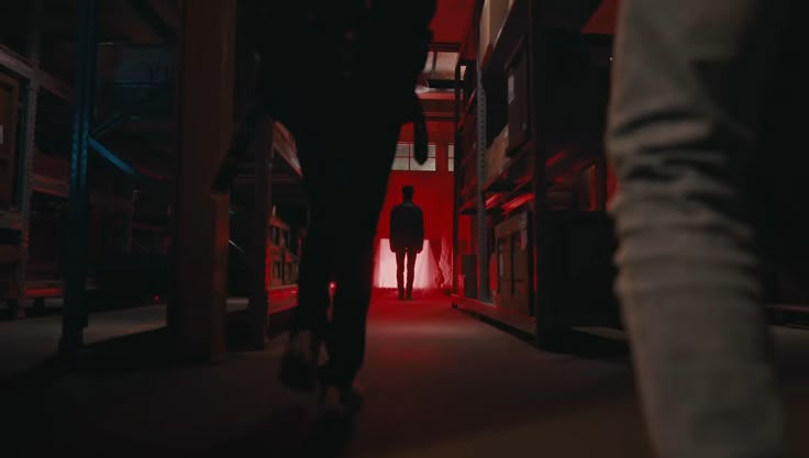 a dimly lit warehouse aisle with shelves of stored items is bathed in a red glow. two blurred figures are walking towards the viewer, while a silhouetted man stands in the distance between the shelves, framed by a window.
