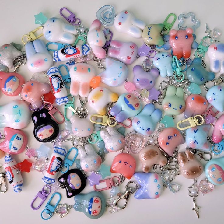 a collection of handmade, kawaiistyle keychains are scattered across a white surface. the keychains feature various designs including bunnies, bears, and other cartoon characterlike shapes. they are made of clay and/or resin and are decorated in pastel colors with painted details such as faces and patterns. many have additional charms like stars and hearts, and are attached to different styles of clips and keyrings. the overall impression is whimsical and colorful, showcasing a collection of cute and unique handmade accessories.