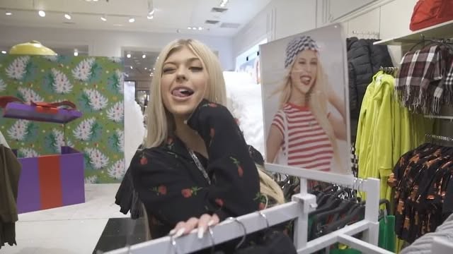 a woman with blonde hair sticking her tongue out is standing in a clothing store in front of a clothing rack. behind her is a poster with a photo of a woman sticking her tongue out, and a display with wrapping paper of christmas trees and a purple and orange present.