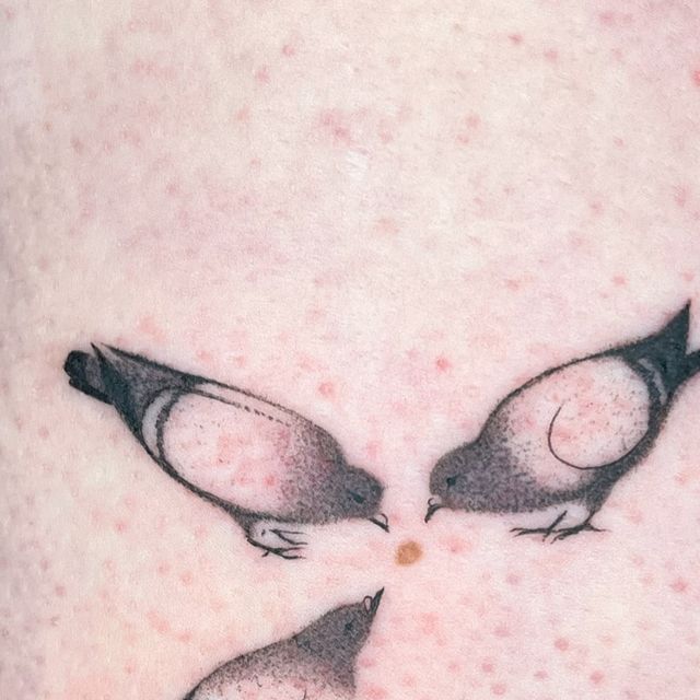 the image shows a realistic tattoo of three pigeons standing around a small circular mark on skin. the pigeons are rendered in detail with gray tones and defined black outlines. the texture of the skin on which the tattoo is placed is visible, showing small red spots.