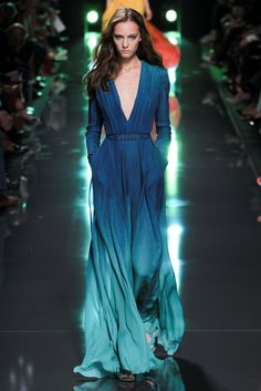 a fashion model walks the runway in a long, flowing blue ombre gown. she is wearing her hair down and has a natural makeup look. the dress is very elegant and looks like it would be perfect for a special occasion. the model's pose and walk are both very confident, adding to the overall allure of the image.