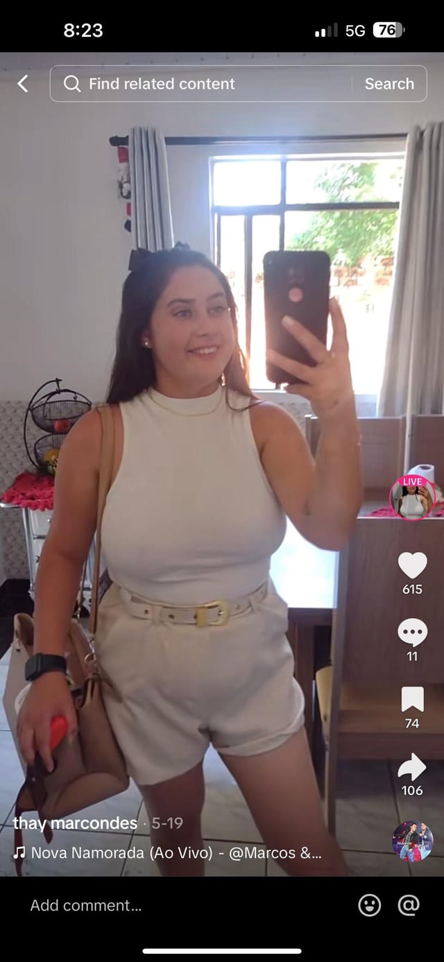a woman takes a selfie with her phone. she's wearing a white shirt, white shorts, and has her dark hair pulled back from her face. she's smiling and looking at the phone. she's posing for a photo in her room.