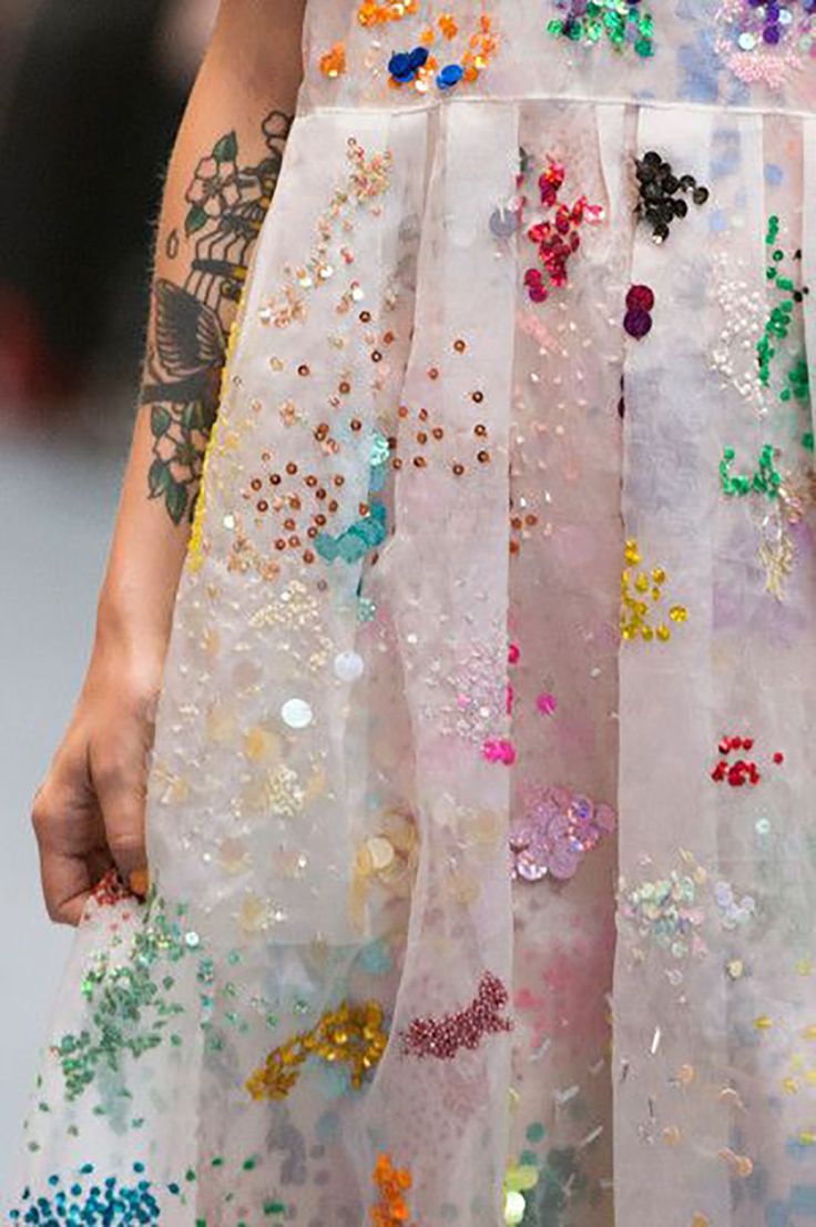 a closeup shot of a woman wearing a white dress with sequins and embroidery. the dress is very detailed and intricate, with a rainbow of colors. the woman's arm is visible, and it has a large tattoo on it. the overall look is very feminine and elegant.