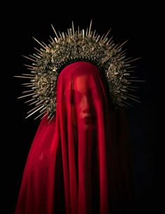 a woman is dressed in a red veil and a crown of thorns. her face is hidden by the veil, but her eyes are visible. the background is black, and the overall image is dramatic and mysterious.