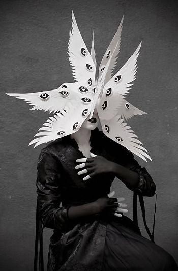 a woman in a black outfit with dramatic makeup is standing in front of a dark backdrop. she is wearing a white paper structure with many cut out eyes covering her face and head. the paper structure looks like wings.  she is striking a pose and looking directly at the viewer, hands gloved and raised to her face.