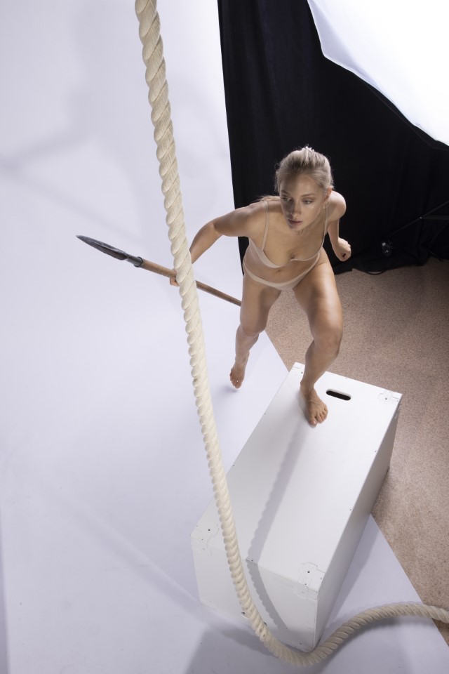 a woman is standing on a white platform in a studio, holding a spear in one hand and gripping a rope with the other, as if climbing. she is barefoot and appears athletic. the background includes a black backdrop and a light source.