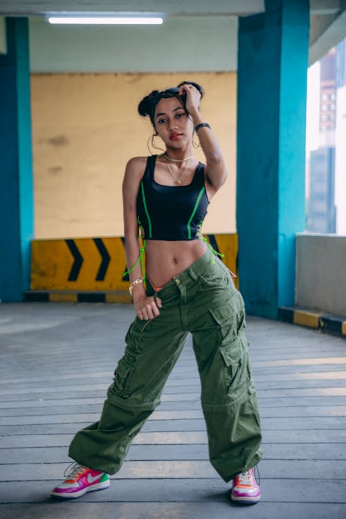 a young asian woman stands confidently in a parking garage, wearing a stylish outfit consisting of olive green cargo pants, a black crop top with lime green accents, and colorful sneakers. she has her hand on her head, and her pose is both relaxed and fashionable. the setting adds an urban and edgy vibe to the overall image.