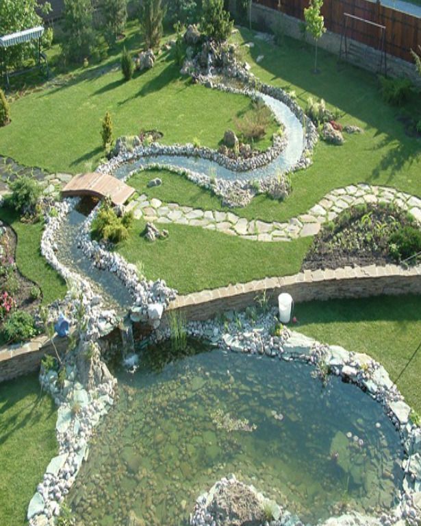 an aerial view of a landscaped backyard with a pond, stream, waterfall, and bridge. the stream winds through the green grass and is lined with stones. the water flows into a pond surrounded by rocks. a small wooden bridge crosses the stream. the landscape includes various plants, trees, shrubs, and flowers. a stone pathway leads through the garden. a white bucket is placed near the pond.