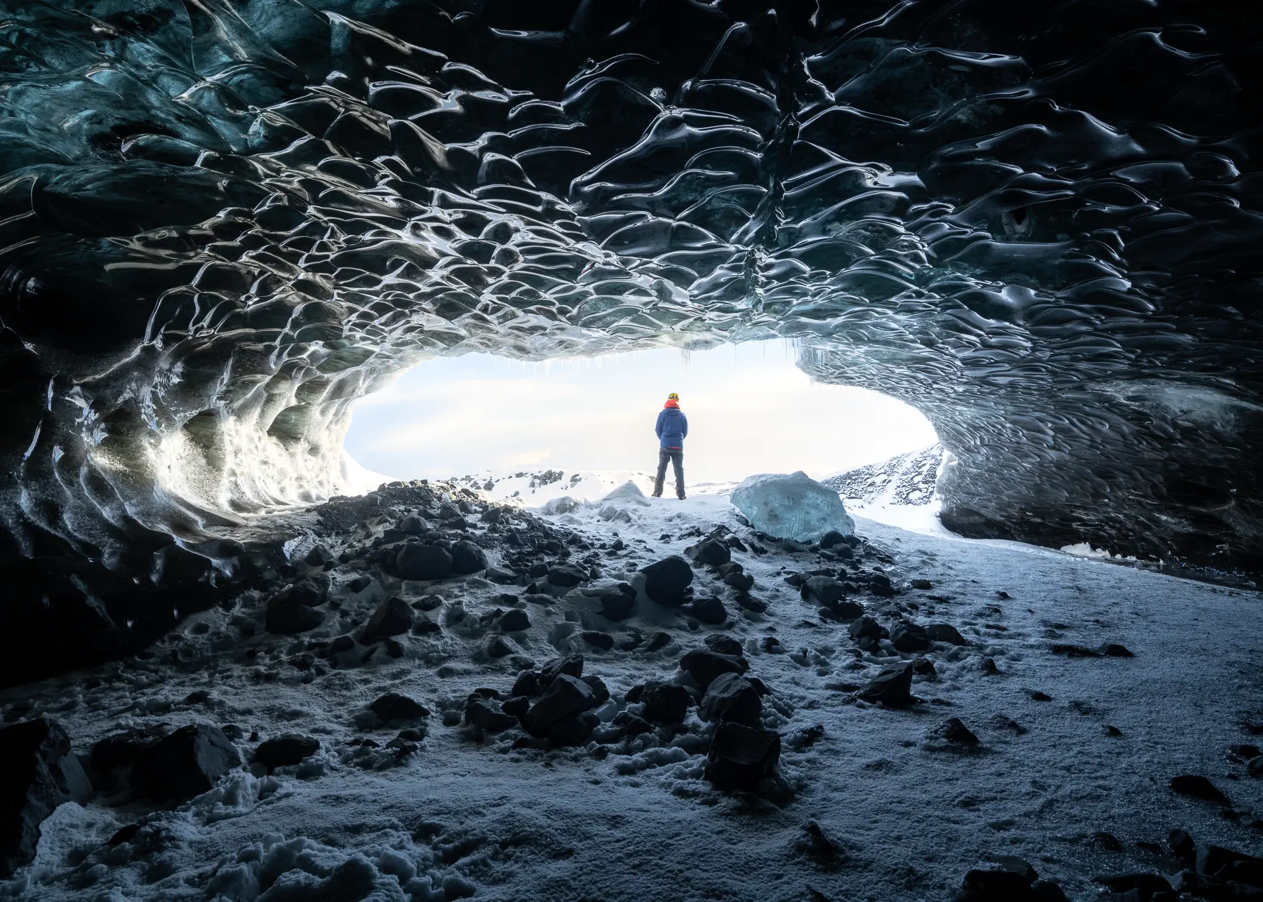 a person stands silhouetted against the light coming from the entrance of a blue ice cave.  the ice is textured and beautiful, with light reflecting off of the ice walls.  the cave is dark inside, with a snowy ground.  the person is wearing a blue jacket, an orange hat, and jeans. they are standing in the center of the cave, looking out at the bright light of the entrance.  the mountains are visible in the distance. the cave has a dark, imposing, yet beautiful aesthetic.