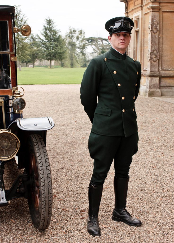 a man in a green uniform, hat, and goggles stands next to a vintage car on a gravel driveway. behind him is a building and trees. he is wearing tall black boots.